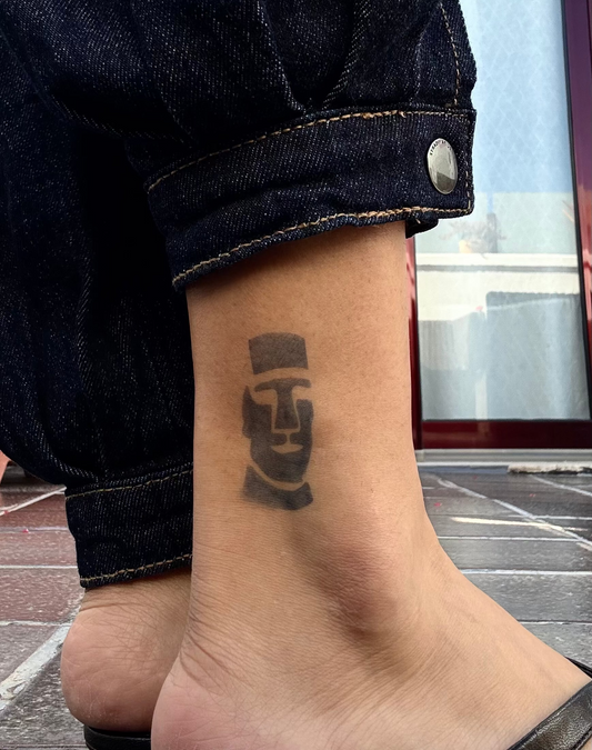 Tatouage temporaire Moai île de Pâques, statue ancestrale polynésienne cheville. Une tête de Moai, statue ancestrale de l'île de Pâques, vue de face. Traits anguleux, front haut, mâchoire massive — aplat noir avec des nuances qui donnent du volume à la pierre. Posée sur la cheville.