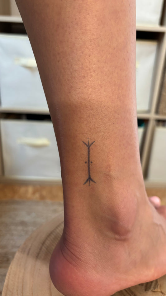 Tatouage temporaire hirondelle amazighe berbère, symbole fidélité retour cheville. Un petit signe amazigh vertical à l'intérieur de la cheville : un axe central traversé d'une fourche en haut et d'une fourche inversée en bas, deux points encadrant le centre. Tracé fin, format minuscule.