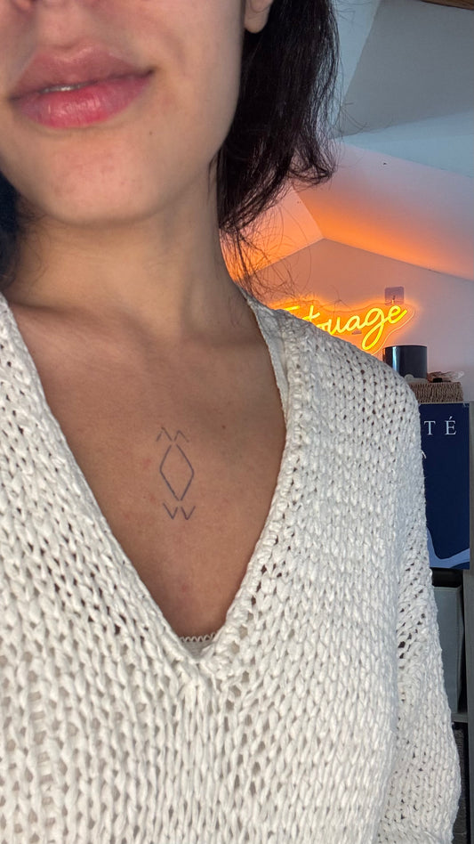 Tatouage temporaire losange amazigh féminin, symbole sacré sterno décolleté. Deux losanges fins empilés à la verticale au creux du décolleté, encadrés en haut et en bas par deux petits chevrons. Tracé léger en fine ligne, posé pile au centre du sterno entre les clavicules.