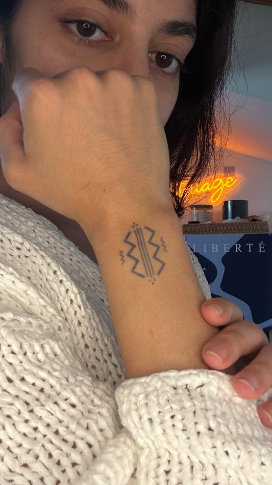 Tatouage temporaire bateau amazigh berbère, glyphe protection sagesse poignet. Un motif amazigh aux lignes tribales : trois bandes verticales centrales encadrées de zigzags symétriques, et quelques points en triangle. Posé sur le poignet, lecture rituelle façon glyphe ancien.
